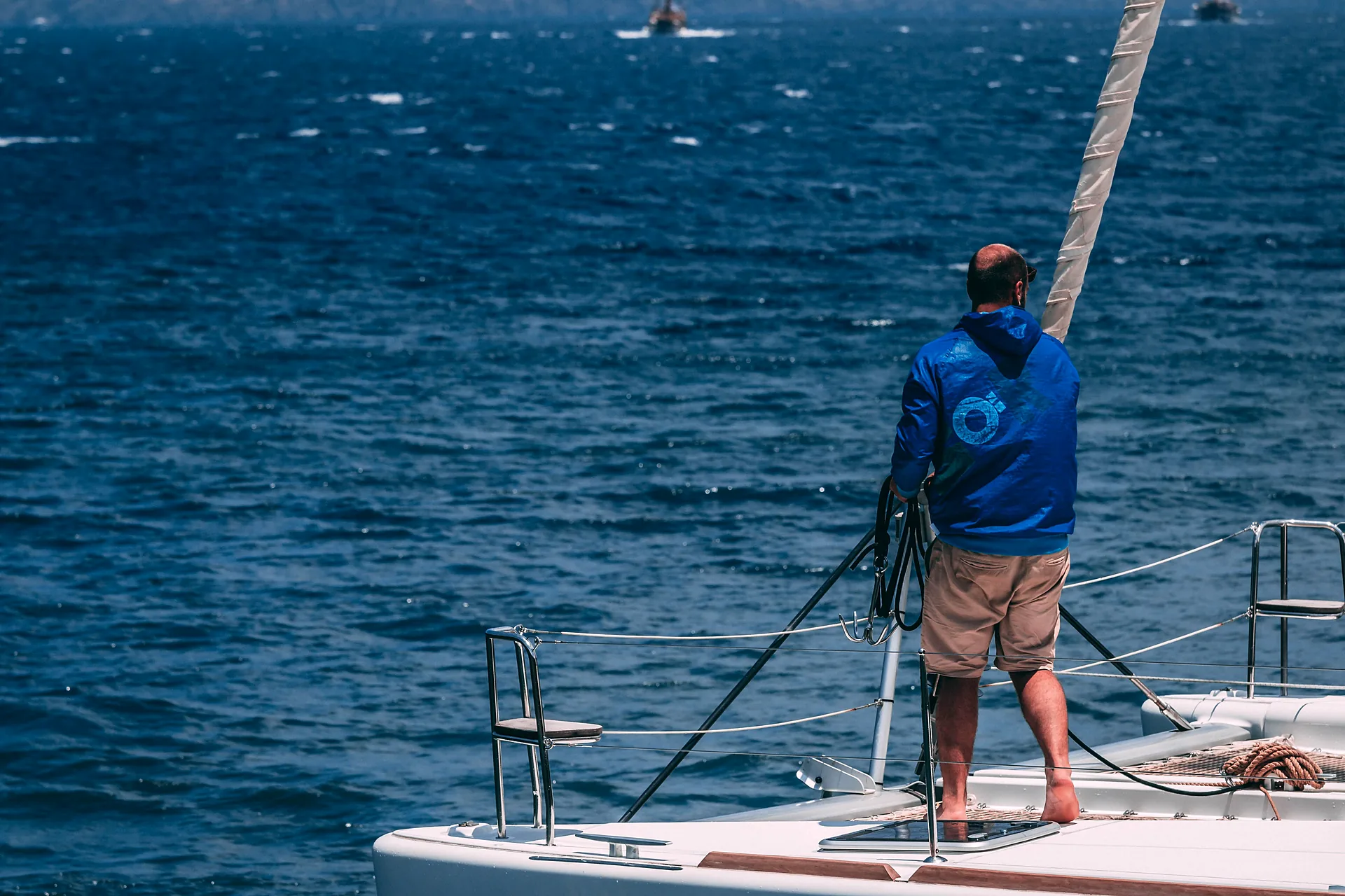 Velerista de pie en la proa de un velero, frente a un mar abierto azul profundo.